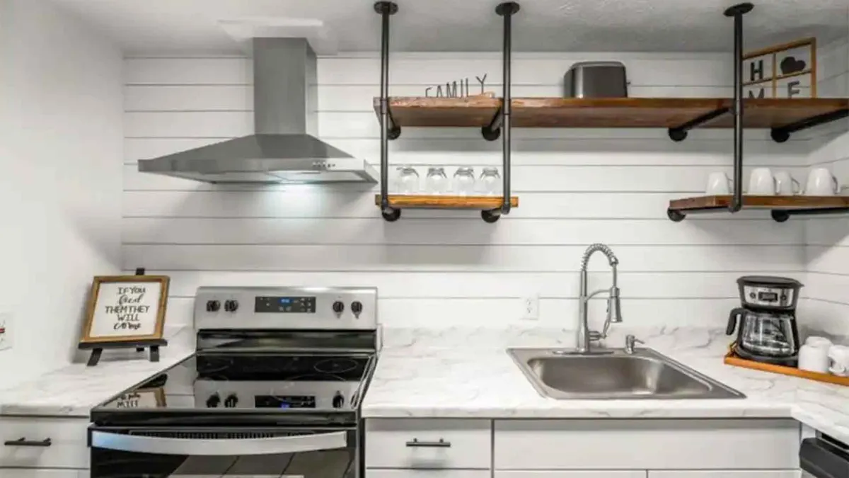 A cozy and inviting kitchen featuring a stainless steel stove, wooden shelves filled with cookware, and a sink, all subtly illuminated by warm natural light. The space feels welcoming and ideal for both everyday meals and festive cooking projects.
