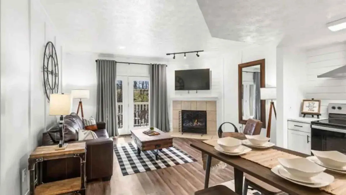 A chic and inviting open-concept living room with a comfortable sofa, coffee table, glowing fireplace, dining table set for meals, and large windows that let in natural light. The space is tastefully decorated with modern accents, emphasizing warmth and relaxation.