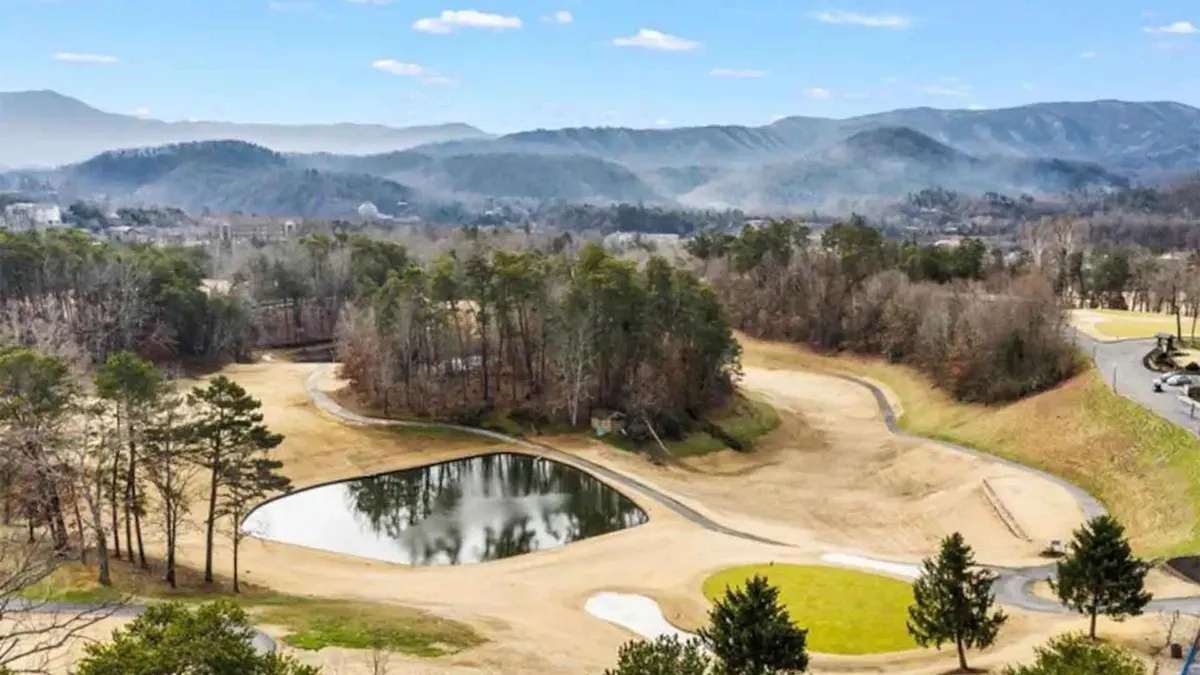 A sweeping resort view featuring a lush golf course, serene pond with morning mist, expansive mountain range, and clusters of trees. The tranquil setting is occasionally animated by golfers and abundant wildlife.