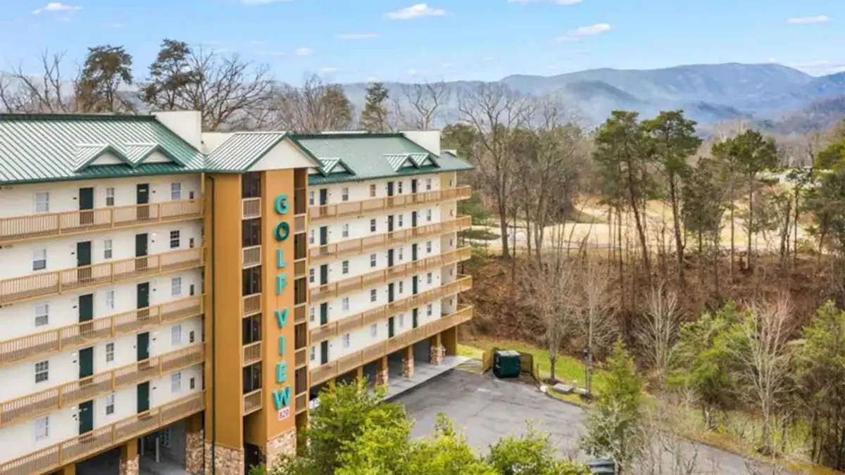 A wide exterior shot featuring the hotel building with balconies overlooking a lush green golf course and a scenic mountain backdrop. A few guests are visible near the course, emphasizing its accessibility and tranquil setting.