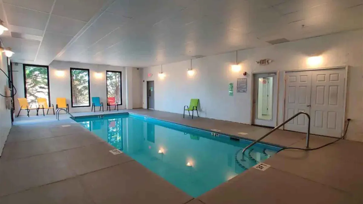 A bright indoor swimming pool area filled with natural light from large windows. Colorful chairs line the poolside, creating a cheerful, inviting atmosphere for kids and adults to enjoy swimming or relaxing after a day of adventure.