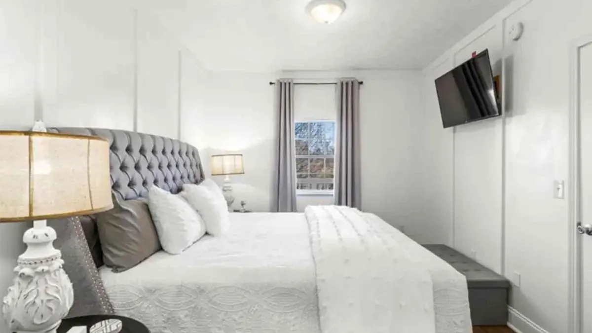 A serene master bedroom featuring a plush bed with a tufted headboard, decorative lamps on bedside tables, a wall-mounted television, and a window with flowing curtains. The room exudes tranquility and privacy, ideal for restful nights.