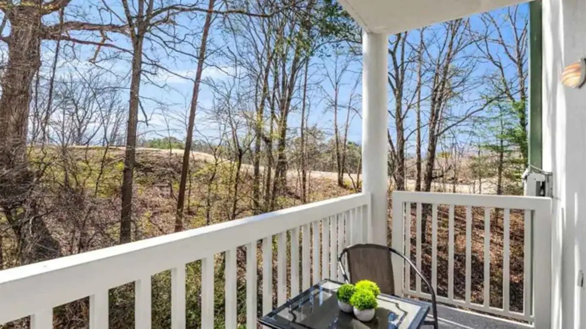 A cozy balcony with a wooden railing and a small table topped with a decorative plant, overlooking a canopy of surrounding trees. The space exudes peacefulness, perfect for quiet reflection or sharing a moment with a loved one.