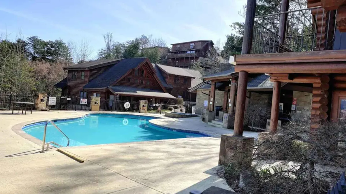 An inviting outdoor swimming pool surrounded by log cabin-style resort buildings and manicured landscaping. Sun loungers are set up for guests to enjoy the sun, and the setting exudes a resort-like, rejuvenating vibe.