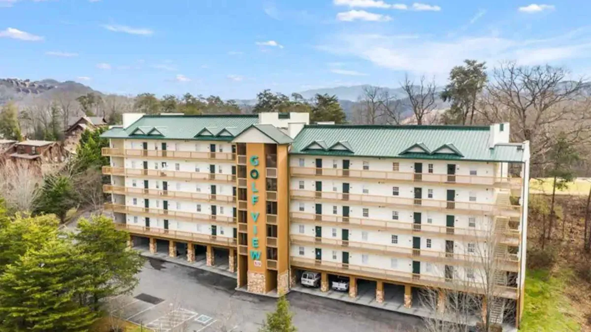 A broad exterior view of the building structure with clear signage, nestled among trees and close to ample parking. The setting is open and inviting, suggesting convenient access to both the condo and nearby resort amenities like golf and local attractions.